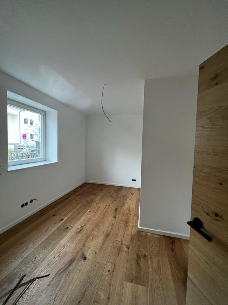 Leerer Raum mit weißen Wänden, hellem Parkettboden, einem Fenster und einem Kabel an der Decke.