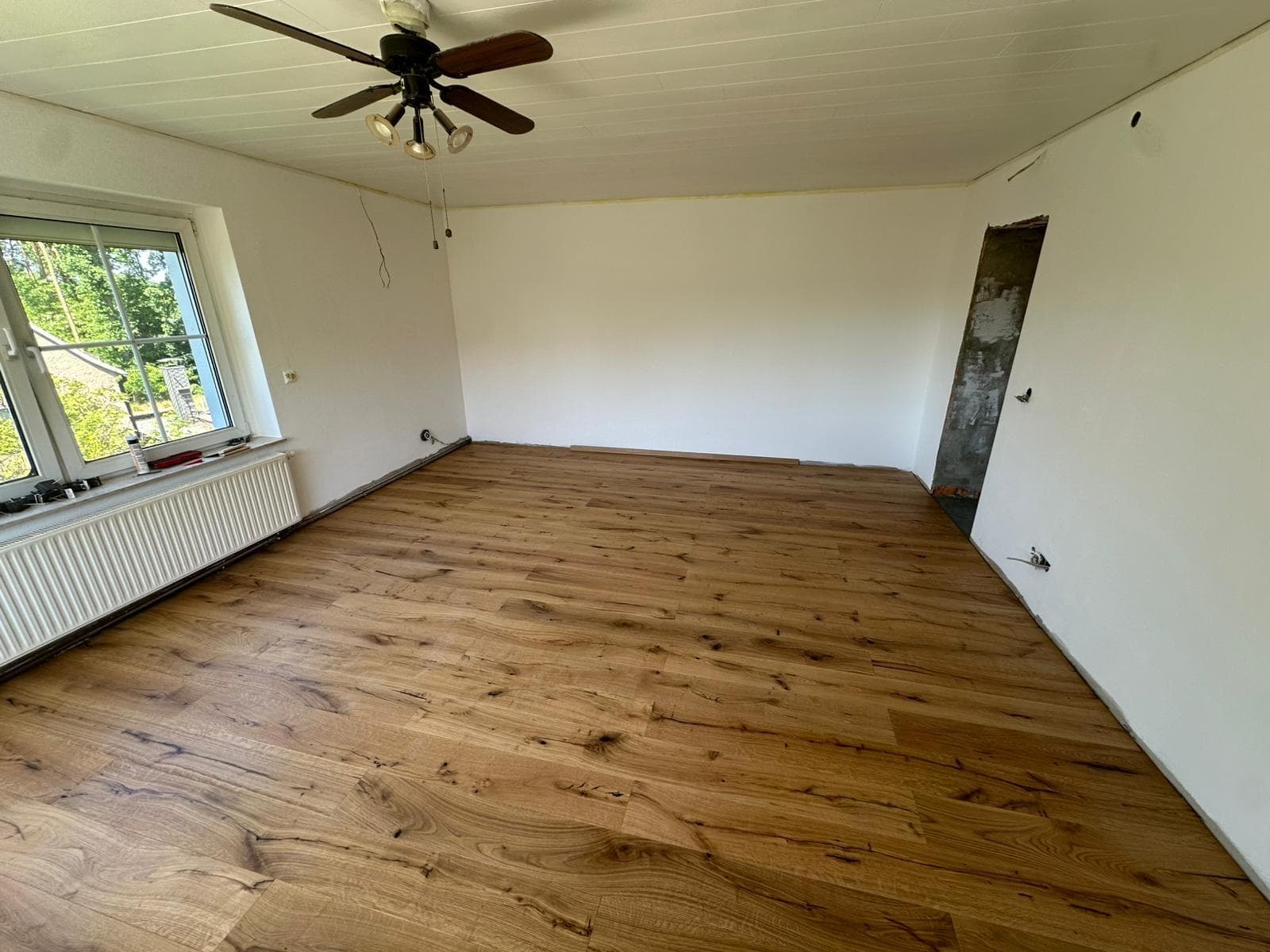 Leerer Raum mit Holzboden, weißen Wänden, Fenster, Heizkörper und Deckenventilator.