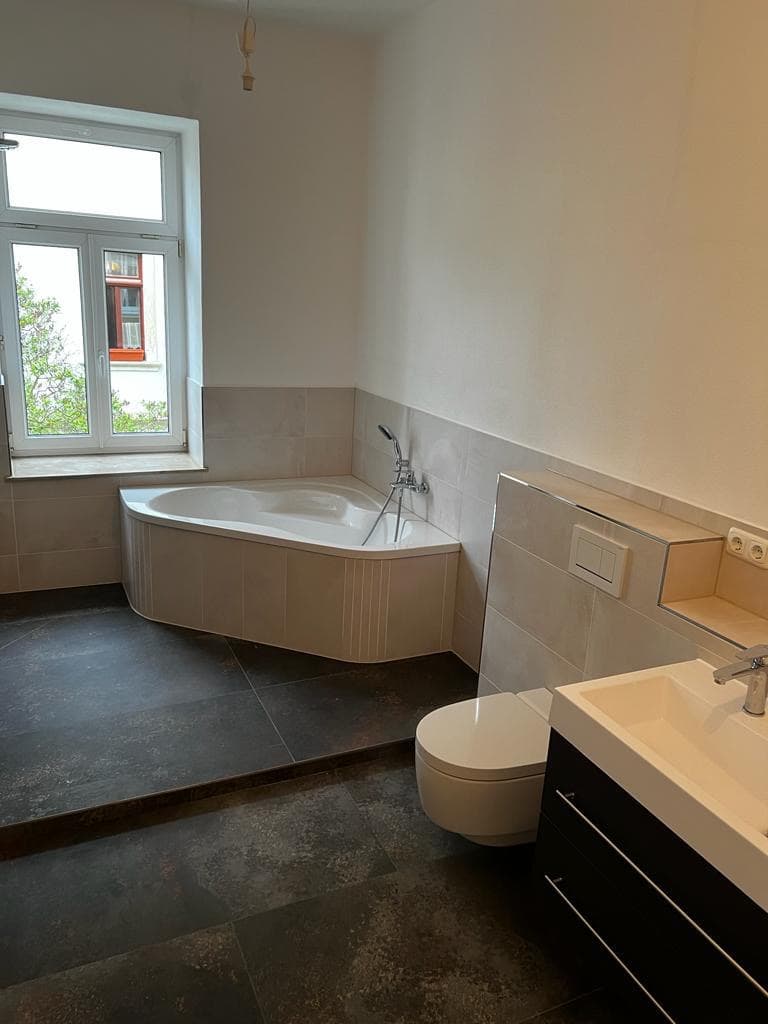 Modernes Badezimmer mit Eckbadewanne, wandhängendem WC, dunklen Bodenfliesen, weißem Waschbecken und großem Fenster.